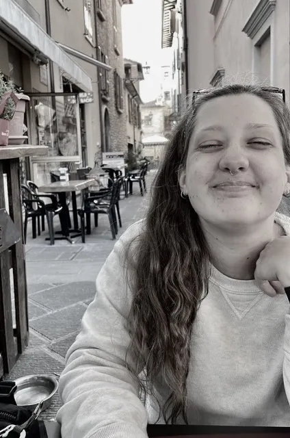 Lynn - a grayscale photo of a long-haired woman in a white crewneck sweatshirt. In the background is an outdoor plaza with stone pavement and small café tables.