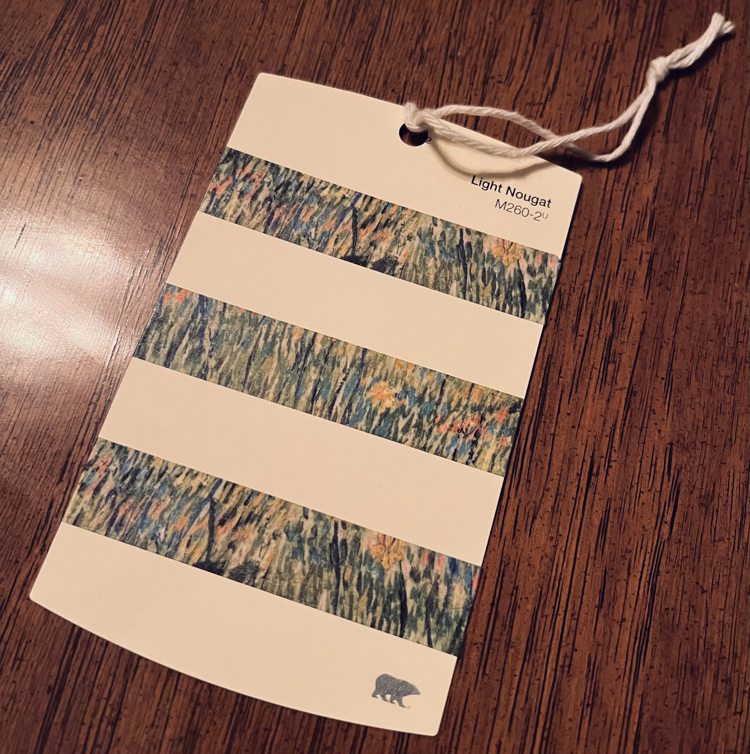 A creamy off-white paint color sample card reading 'Light Nougat // M260-2u'. Broad horizontal stripes on its surface are formed by washi tape printed with Van Gogh's "Patch of Grass" impressionist painting -- vibrant green, blue and pale pink upward brushstrokes with yellow flowers dotted throughout.